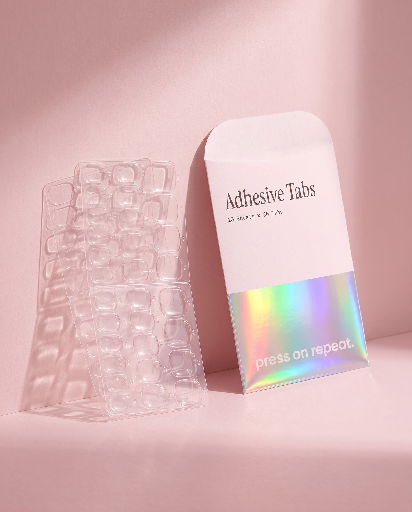 Sheets of clear adhesive nail tabs standing upright against the holographic Press On Repeat envelope.