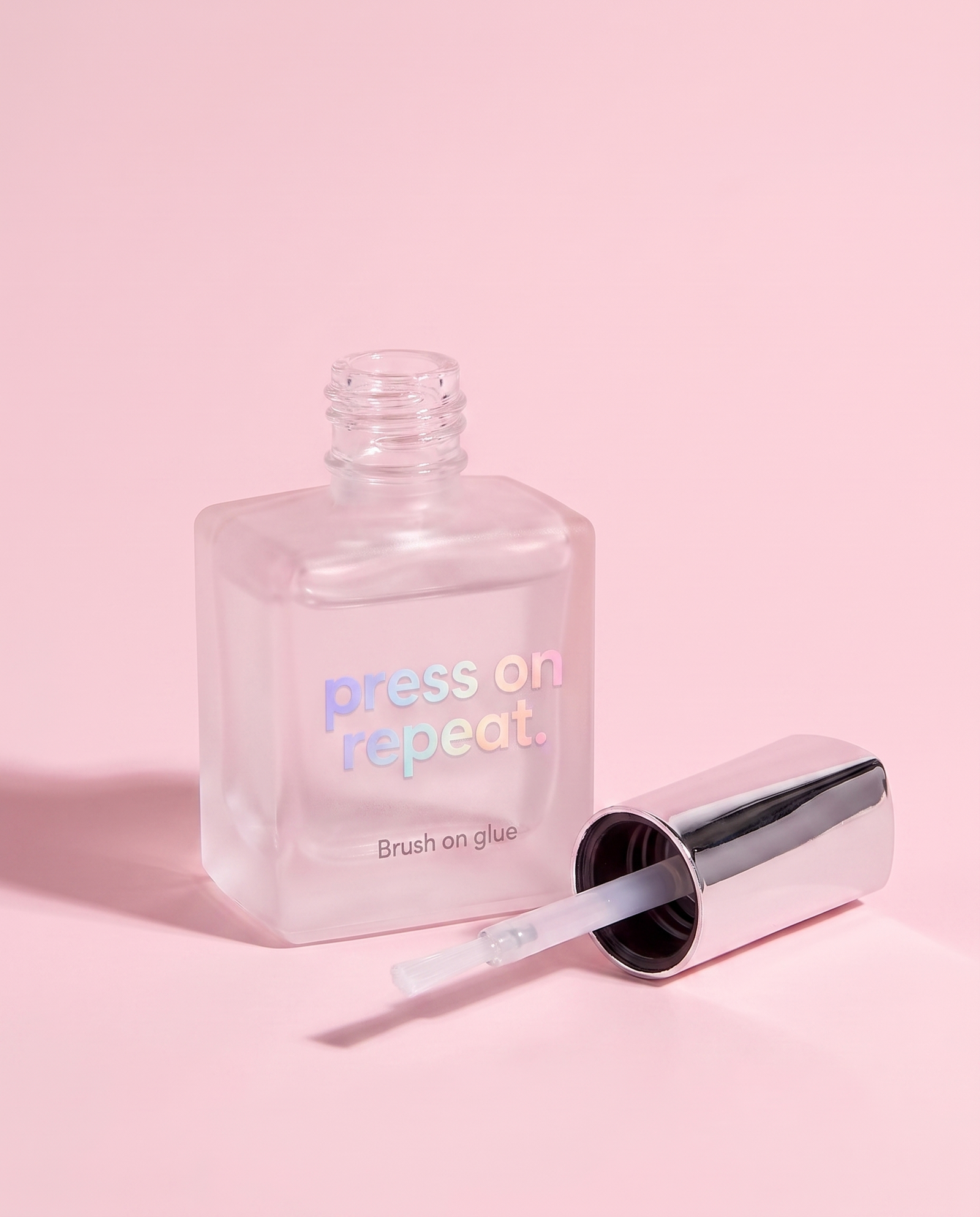 Brush-on nail glue open at an angle, chrome cap and brush lying beside the bottle.