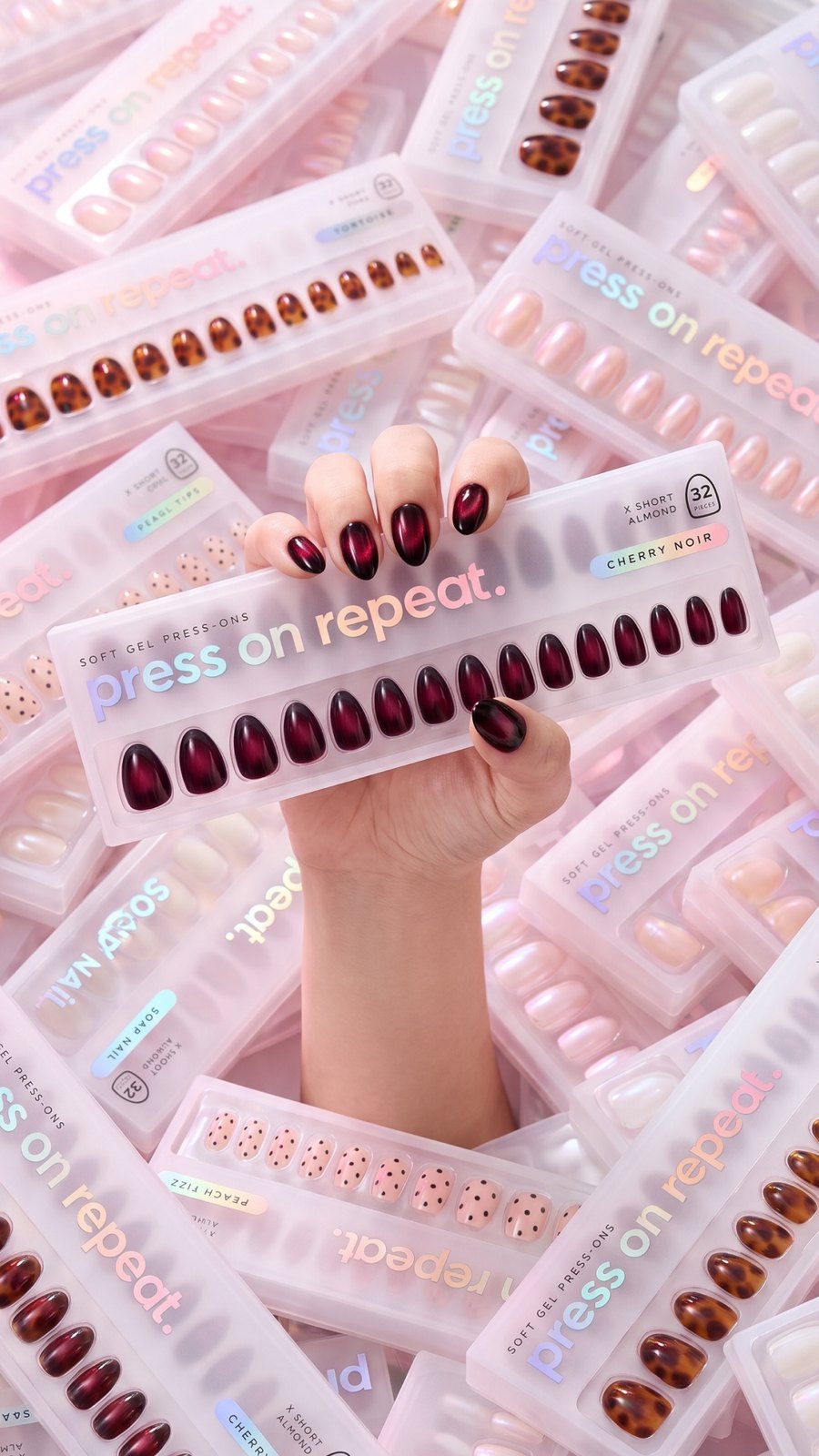 A hand wearing dark red press-ons reaching out of a pile of Press On Repeat packs.